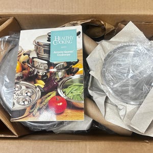 May include: A cardboard box containing stainless steel cookware pieces wrapped in protective paper and plastic, accompanied by a cookbook titled Healthy Cooking with Amway Queen Cookware featuring images of stacked pots and fresh produce.