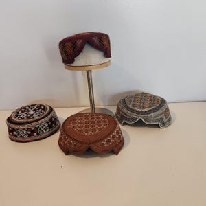 Peut inclure: Quatre calottes rondes avec des broderies complexes. Une calotte bordeaux est posée sur un support en métal. Trois autres sont posées sur une surface : une noire à motifs floraux blancs, une marron à coutures géométriques et une grise à motifs sombres.