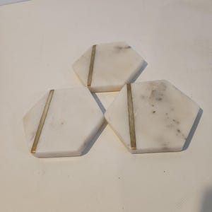 May include: Three hexagonal white marble coasters with natural grey veining. Each coaster features a single, straight, vertical brass-colored metal inlay strip running off-center across the surface of the stone, creating a geometric design.