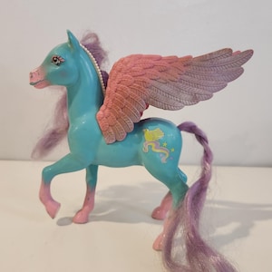 Puede incluir: Un juguete de pegaso de plástico turquesa con grandes alas rosas texturizadas. La figura presenta una crin y cola moradas, cascos rosas y un gráfico de un pájaro amarillo con pequeñas estrellas en el flanco. Tiene ojos pintados.