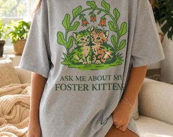 Puede incluir: Una camiseta gris jaspeado con un gráfico de dos gatitos sentados bajo un arco de hojas verdes con flores naranjas. El texto debajo de los gatitos dice "ASK ME ABOUT MY FOSTER KITTENS" en letras mayúsculas de color verde oscuro.