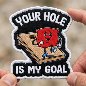 May include: An embroidered patch with a black background and white border featuring a tan cornhole board and a red square character with a face, arms, and blue shoes. The text reads YOUR HOLE IS MY GOAL in white capital letters on the black background.