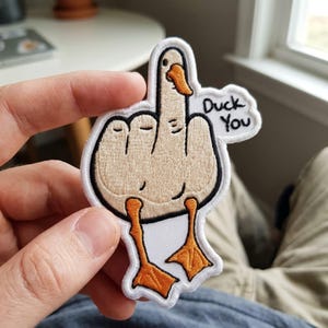 May include: An embroidered patch featuring a light tan duck with an orange beak and feet. The duck is depicted with its wing raised in a middle finger gesture. A white speech bubble attached to the side contains the black embroidered text Duck You.