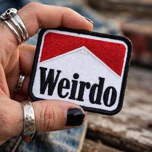 May include: A rectangular embroidered patch featuring a white background with a red chevron design at the top. The word "Weirdo" is stitched in bold black letters across the center. The patch has a thick black stitched border around the edges.