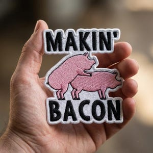 May include: An embroidered patch with a white border featuring two pink pigs and the black text MAKIN' BACON. The design shows one pig standing behind another. The patch has a white background and is shaped to follow the outline of the text and pigs.