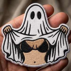 May include: An embroidered patch featuring a white ghost sheet with black oval eyes. The ghost is lifting the bottom of the sheet to reveal a tan-colored torso wearing a black bikini top. The design has a white border and black stitching details.