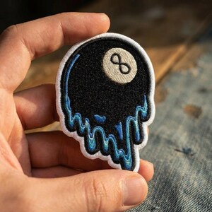 May include: An embroidered patch shaped like a black billiard ball with a white number eight circle. The bottom edge features a blue and black melting or dripping effect with a white border surrounding the entire design on a fabric base.