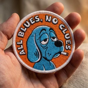 May include: A circular embroidered patch featuring a blue dog with droopy eyes and a cigarette in its mouth, set against an orange background. The text "ALL BLUES, NO CLUES" is stitched in white along the top edge, bordered by a white stitched rim.