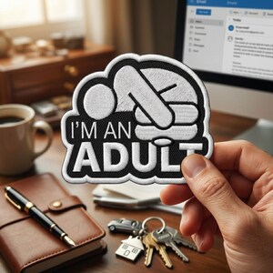 May include: A black and white embroidered patch featuring a stick figure lying face down on a desk with the text I'M AN ADULT printed in bold white capital letters below the figure. The patch has a thick black border and a white background.