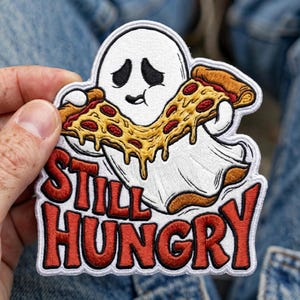 May include: An embroidered patch featuring a white ghost with a sad expression holding a large slice of pepperoni pizza with melted cheese. Below the ghost, the text STILL HUNGRY is stitched in bold red letters with a black outline on a white background.