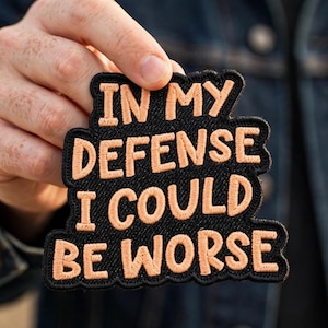 May include: A black embroidered patch with a scalloped edge featuring the peach-colored text "IN MY DEFENSE I COULD BE WORSE" in a bold, sans-serif font. The patch has a textured fabric surface and is shaped to follow the contour of the text.
