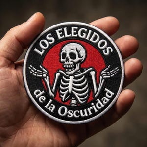 May include: A circular embroidered patch featuring a white skeleton with arms raised in a shrug gesture against a red circular background. The black border contains the white text LOS ELEGIDOS de la Oscuridad in a bold, capitalized font.