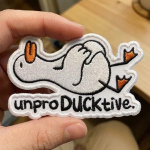 May include: An embroidered patch featuring a white duck lying on its back with orange feet and an orange beak. The duck has black outlines and is positioned above the black embroidered text that reads, "unproDUCKtive." with a period at the end.