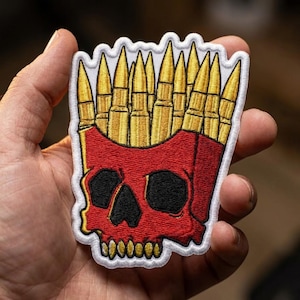 May include: An embroidered patch featuring a red skull shaped like a french fry container, with gold bullet casings standing upright inside like french fries. The design has a white border and black stitching for the eye sockets and nasal cavity.