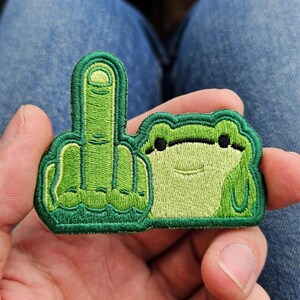 May include: An embroidered patch featuring a green frog with a light green belly and dark eyes, positioned next to a green hand gesture showing a middle finger. The design uses various shades of green thread with a dark green border outline.
