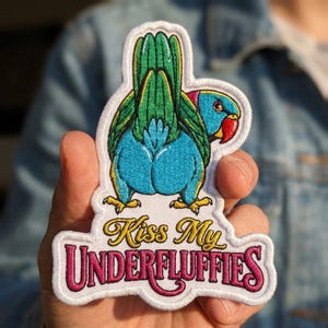 May include: An embroidered patch featuring a blue and green parrot viewed from behind, with its head turned to the side. The parrot has a red beak and yellow feet. Below the bird, the text reads "Kiss My UNDERFLUFFIES" in gold and pink.