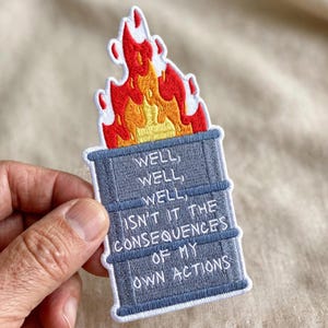 May include: An embroidered patch featuring a grey barrel engulfed in bright red, orange, and yellow flames. The barrel has white text stitched on the front that reads: WELL, WELL, WELL, ISN'T IT THE CONSEQUENCES OF MY OWN ACTIONS.