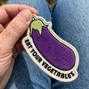 May include: An embroidered patch shaped like an eggplant with a purple body and light green stem. The patch features a cream-colored border and the black embroidered text EAT YOUR VEGETABLES curved along the bottom edge of the vegetable shape.