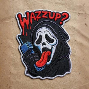 May include: An embroidered patch featuring a black-hooded figure with a white ghost mask, holding a blue telephone handset. The figure has a long red tongue hanging out. Above the figure, the text WAZZUP? is stitched in red with a dripping effect.