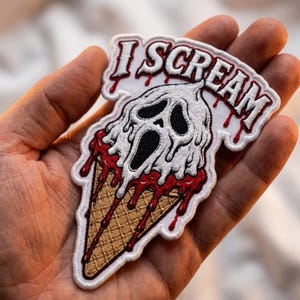 May include: An embroidered patch featuring a tan waffle cone topped with a white, melting ghost-like figure with black eye and mouth cutouts. Red liquid drips down the cone, and the text I SCREAM is embroidered in white with red outlines.