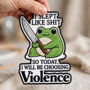 May include: An embroidered patch featuring a green frog holding a silver knife with a brown handle. The text reads I SLEPT LIKE SHIT SO TODAY, I WILL BE CHOOSING VIOLENCE. The patch has a white background, black border, and detailed stitching.
