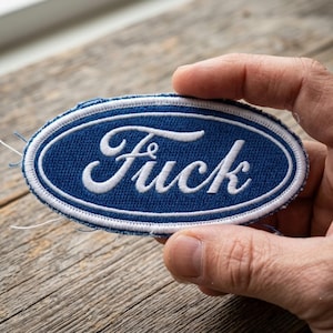 May include: An oval-shaped embroidered patch featuring a blue background with a white border and the word "Fuck" stitched in white script lettering. The design mimics a classic automotive logo style with clean, white thread embroidery.