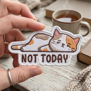 May include: An embroidered patch featuring a white, brown, and tan calico cat lying down with a tired expression. The cat has pink inner ears and brown outlines. Below the cat, the text NOT TODAY is embroidered in brown capital letters.