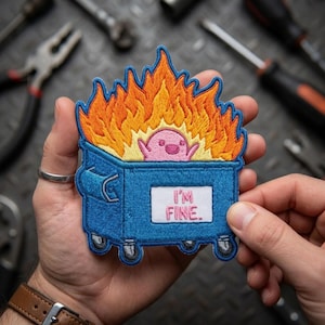 May include: An embroidered patch featuring a blue dumpster engulfed in orange and yellow flames. A small pink creature with a simple face sits inside the dumpster. A white rectangular sign on the front of the dumpster displays the text I'M FINE.