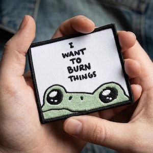 May include: A square embroidered patch with a white background and a black border. The text I WANT TO BURN THINGS is stitched in black letters above a light green frog face with large black eyes and white highlights at the bottom of the patch.