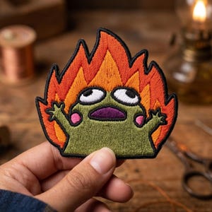 May include: An embroidered patch featuring a green frog character with wide eyes and pink cheeks, surrounded by stylized orange and red flames. The design is outlined in black thread, creating a bold contrast against the vibrant colors of the fire.