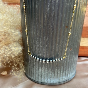 May include: A delicate gold chain necklace featuring a central row of small, polished dark grey metallic beads. The necklace is constructed with a fine gold link chain that includes evenly spaced gold beads along the sides, leading to the center.