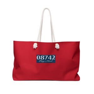 May include: A bright red tote bag featuring white rope handles secured with metal grommets. Centered on the front is a dark blue rectangular patch displaying the white text 08742 and the script word Bay Head below the numbers.