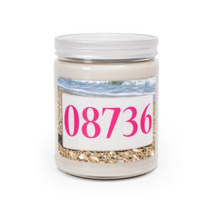 Könnte beinhalten: Eine Kerze im klaren Glasgefäß mit cremeweißem Wachs und silbernem Deckel. Das Etikett zeigt eine Strandszene mit Sand und Meereswellen sowie die leuchtend pinken Ziffern 08736 auf einem weißen, rechteckigen Hintergrund.