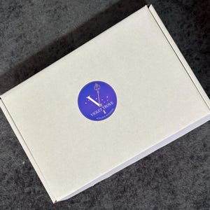 May include: A rectangular white cardboard shipping box featuring a circular sticker in the center. The sticker has a blue to purple gradient background and displays a gold key icon above the text VIOLET TROVE and UNLOCK WONDER.