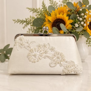 May include: An ivory satin clutch bag featuring a silver-toned metal kiss-lock closure. The front of the bag is decorated with an asymmetrical floral lace appliqué embellished with small white beads and sequins arranged in a delicate pattern.