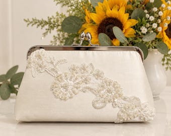 May include: An ivory satin clutch bag featuring a silver-toned metal kiss-lock closure. The front of the bag is decorated with an asymmetrical floral lace appliqué embellished with small white beads and sequins arranged in a delicate pattern.