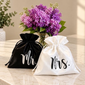 May include: Two satin drawstring pouches, one black with silver glitter text reading Mr. and one white with black glitter text reading Mrs. The pouches are positioned side by side, featuring a smooth, shiny fabric finish and matching drawstrings.