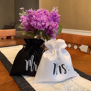 May include: Two satin drawstring bags, one black and one white, sit on a table runner. The black bag has "mr" in white script, and the white bag has "mrs" in black script. A bouquet of purple flowers is in the background.