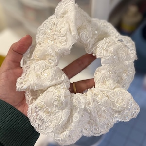 May include: Ivory lace scrunchie with delicate floral patterns and scattered pearl beads. The scrunchie is a hair accessory, perfect for adding a touch of elegance to any hairstyle. The lace is soft and the pearls add a subtle shimmer.