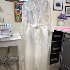 May include: Ivory-colored satin robe displayed on a mannequin. The robe features long sleeves, a tie belt, and delicate embroidered detailing on the upper chest. The robe is knee-length and has a smooth, lustrous finish, suggesting a luxurious feel.