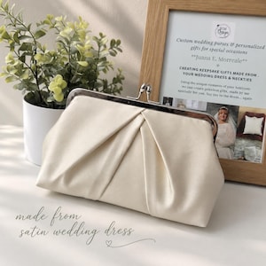 May include: An ivory satin clutch purse featuring a silver-toned metal frame with a kiss-lock closure. The exterior fabric is gathered into soft pleats across the front, creating a structured yet elegant appearance for this formal accessory.