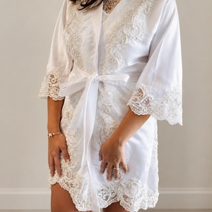 May include: A white satin robe featuring intricate lace trim along the front opening, neckline, and wide sleeves. The lace is embellished with small white beads and sequins. The robe includes a matching white satin tie belt at the waist.