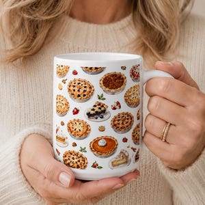 May include: A white ceramic mug featuring a repeating pattern of various baked pies, including fruit-filled pies with lattice crusts, pumpkin pies with whipped cream, and individual slices of pie, interspersed with small strawberries and leaves.