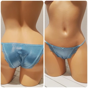 Puede incluir: Un par de bragas estilo bikini de color azul claro con un tejido brillante de acabado metálico en los paneles delantero y trasero. La prenda está decorada con un delicado encaje en los bordes y un pequeño lazo en el centro de la parte delantera.