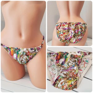 May include: Underwear featuring a vibrant, multi-colored marbled pattern with swirls of pink, green, yellow, and purple. The garment has a thin waistband, a small white bow at the center front, and elasticized leg openings with a ruffled edge finish.