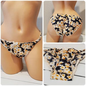 May include: Bikini-style underwear featuring a black background with a white and yellow daisy floral pattern. The garment includes a small white bow at the center front waistband and a delicate white scalloped trim along the edges.