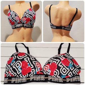 May include: A black and white bralette featuring a repeating logo pattern and red rose floral print. The garment includes black adjustable shoulder straps, a wide elastic band with the text PINK in white, and a hook-and-eye closure at the back.