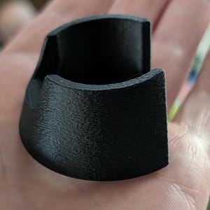 Op de afbeelding: Een kleine, zwarte, C-vormige component met een getextureerde, gelaagde oppervlakteafwerking. Het object heeft een gebogen buitenwand en een holle binnenkant, wat een 3D-geprinte constructie met zichtbare horizontale lijnen laat zien.