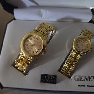 Può includere: Un set di due orologi al quarzo Geneva color oro con cinturini a maglie abbinati, presentati in una scatola bianca e nera. La scatola riporta la scritta Geneva Classic Collection e un cartello con un R.R.P. di 149,99 $.