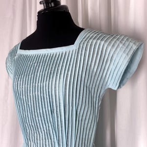 May include: A light blue dress featuring a square neckline and short sleeves. The bodice is constructed with vertical pleats that run from the neckline to the waist, creating a textured appearance across the front of the garment.